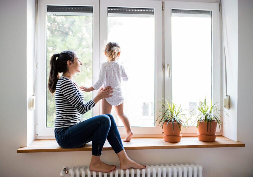 Mother with her little daughter looking out of window Beautiful young mother sitting on window sill with her cute little daughter looking out of window